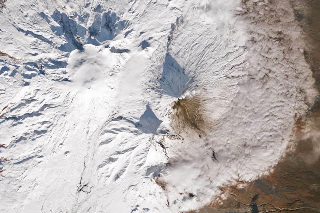 NASAのインスタグラム：「#Landsat 8 captured two neighboring volcanoes puffing away on Russia’s ever-fiery Kamchatka Peninsula in mid-October. Kamchatka Peninsula is a land of volcanoes, home to more than 300 of them.  Image description: Satellite image of the Kamchatka Peninsula. The landscape is powdery white, with a rocky texture. Near the center of the image are two volcanoes releasing white plumes of smoke. The peaks cast shadows on the snow as the Sun traversed low in the autumn sky.」