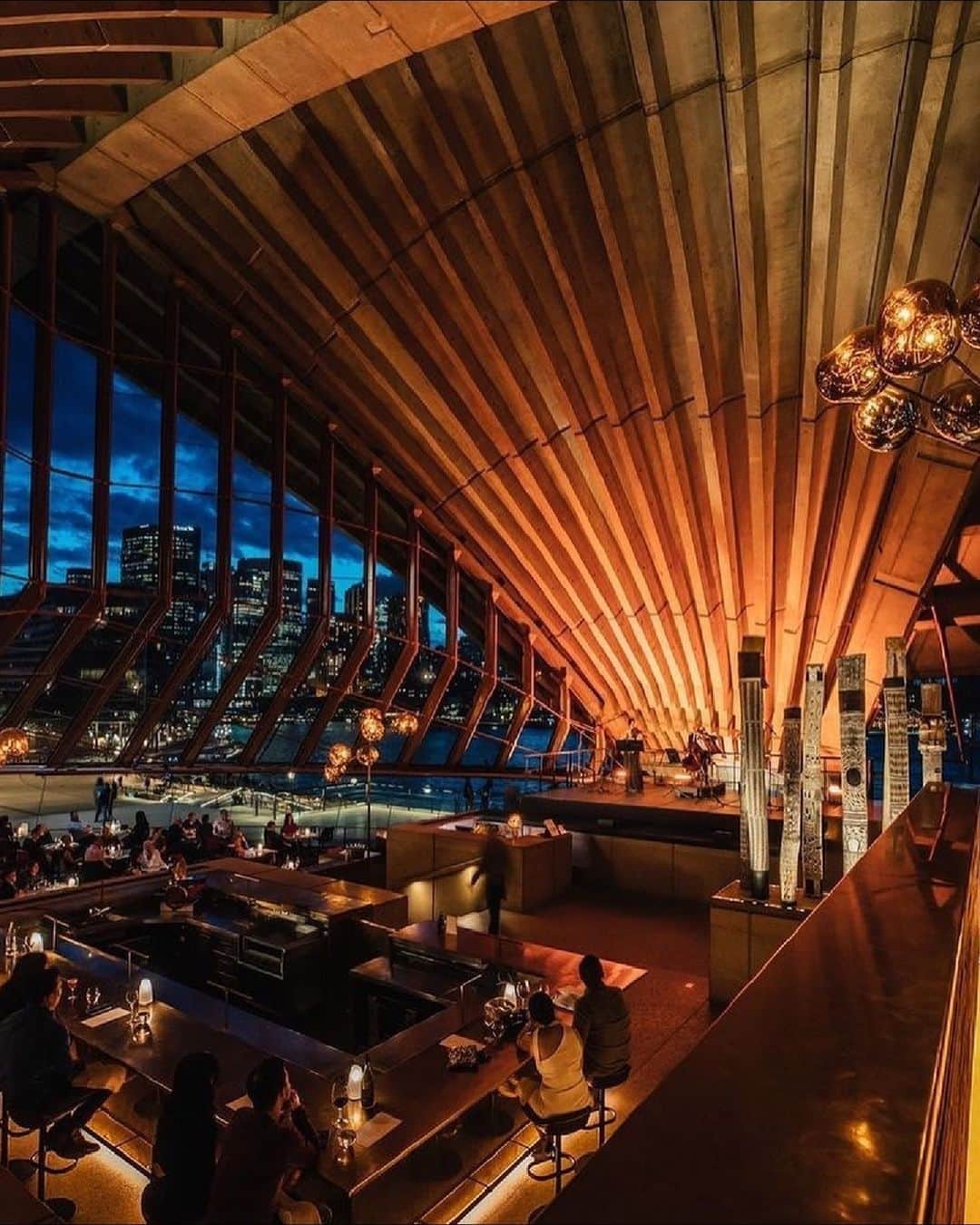 Australiaのインスタグラム：「It's not every day you get to dine beneath the sails of the @sydneyoperahouse 🤩 Step inside @bennelong_sydney, the acclaimed @sydney restaurant by @chefpetergilmore. Overlooking Tubowgule (#SydneyHarbour) with unbeatable views of the #SydneyHarbourBridge, this hatted restaurant at the #SydneyOperaHouse has several semi-private dining spaces for groups of 10-28 guests. Alternatively, the whole venue can be hired exclusively for up to 100 guests seated or 300 for a cocktail-style event.   #ComeAndSayGday #SeeAustralia #MeetInAus #FeelNSW #FeelNewSydney  ID: The interior of a restaurant. Dinners enjoy their meals under warm, moody lighting. Angled walls arch up towards the ceiling, as glass windows show a peak of a city skyline.」