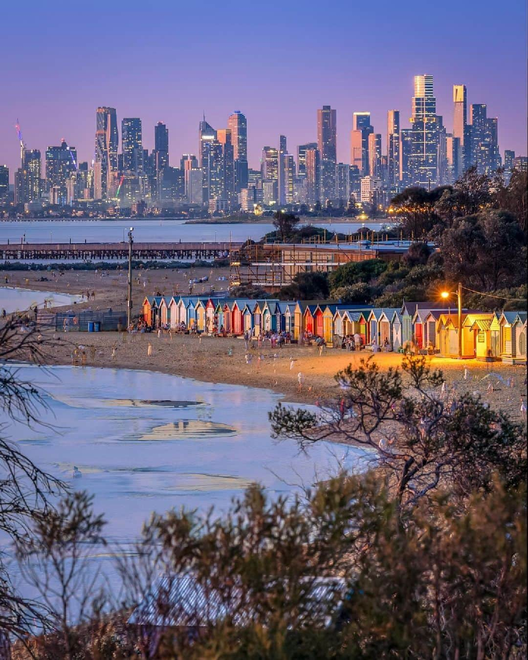 Australiaのインスタグラム：「Naarm (@visitmelbourne) looks perfect from every angle 💕Captured here by @a.j.wilko, it's impossible not to feel happy at the sight of the brightly painted #BrightonBeach bathing boxes - an ode to lazy summer days on #Victoria's sandy coast 🏖️ Try checking out @brightonbathshealthclub here, where a private beach and ocean pool await, then head to @baysidegallery for quirky exhibitions from talented local artists 🖼️   #SeeAustralia #ComeAndSayGday #VisitVictoria #VisitMelbourne  ID: Peering through a leafy tree towards a row of brightly coloured bathing boxes along the coastline, with a city skyline in the distance during the final moments of sunset.」