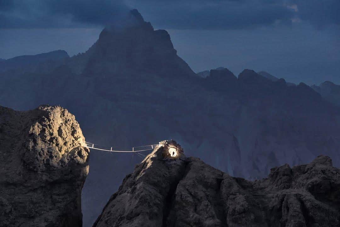 thephotosocietyのインスタグラム：「Photo by Robbie Shone @shonephoto | The longest suspension bridge in the Dolomites is part of the Ivano Dibona via ferrata, a restored historical route that was once used by Italian troops during World War I. Mountains always remind us how small we really are, and this is especially true in the Dolomites. As climbers and mountaineers, we’re always looking for perfect blue skies, but these mountains often look their best with some clouds breaking up the panorama while the sun rapidly disappears. As the peaks in the distance begin to recede into darkness and the shafts of light become even thinner and weaker, the already extraordinary ambience of this fascinating and beautiful place becomes, for a fleeting moment, even more haunting.」