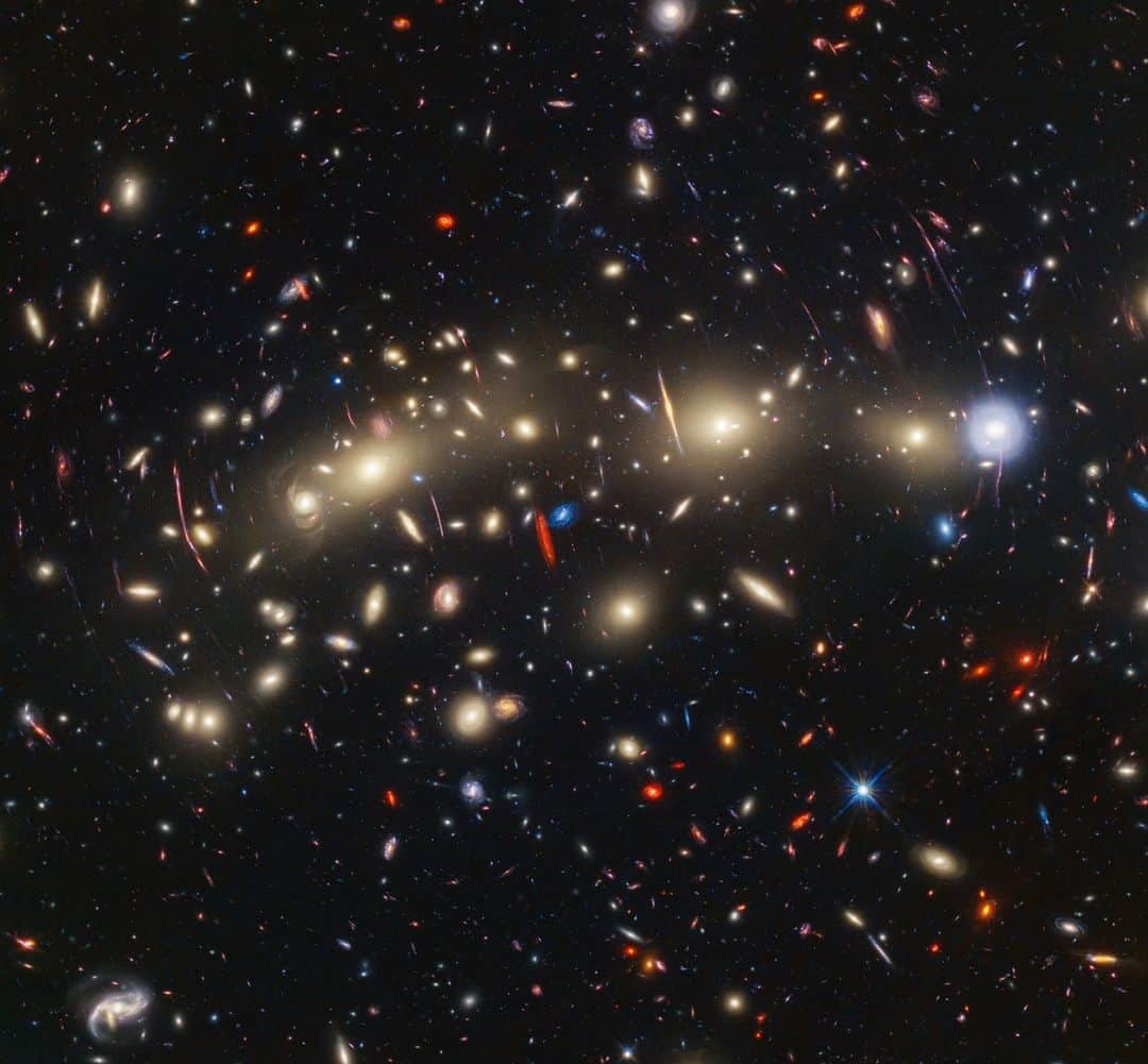 NASAのインスタグラム：「That rainbow connection 🌈 @NASAHubble and @NASAWebb teamed up for this image of giant galaxy cluster MACS0416, one of the most colorful views of the universe. Their views combine visible and infrared light, revealing galaxies hidden in visible alone.  Image description: A field of galaxies on the black background of space. In the middle, stretching from left to right, is a collection of dozens of yellowish spiral and elliptical galaxies that form a foreground galaxy cluster. They form a rough, flat line along the center. Among them are distorted linear features, which mostly appear to follow invisible concentric circles curving around the center of the image. The linear features are created when the light of a background galaxy is bent and magnified through gravitational lensing. At center left, a particularly prominent example stretches vertically about three times the length of a nearby galaxy. A variety of brightly colored, red and blue galaxies of various shapes are scattered across the image, making it feel densely populated. Near the center are two tiny galaxies compared to the galaxy cluster: a very red edge-on spiral and a very blue face-on spiral, which provide a striking color contrast.  #Space #NASA #Hubble #Webb #Galaxy」