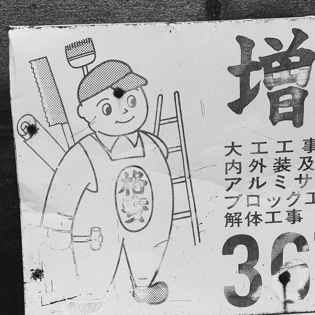 武井義明のインスタグラム：「バス停でもない、交通量も多くない丁字路の歩道角にある広告入りベンチ。誰かが座ってるところは見たことがないけど、坂の途中だから休憩する人もいるのかな。広告は昭和っぽい味わい。「格安」って描かれたオーバーオールのデザインは攻めてますね。ジュンヤで売ってたら買うかも。あと道具は仕舞った方が安全だと思うよ！」