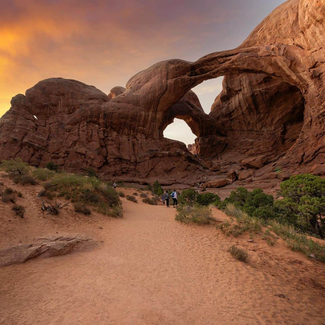 アメリカ内務省のインスタグラム：「It’s National Hiking Day! 🥾    Hiking is a great way to see wildlife and bask in nature’s beauty. Hike alone for peace and clarity or with friends for a fun bonding experience.    Photo at @archesnps by Ricky Barnard    #publiclands #hiking #archesnps #utah    Alt Text: The sun sets behind Double Arch with a few people standing nearby.」